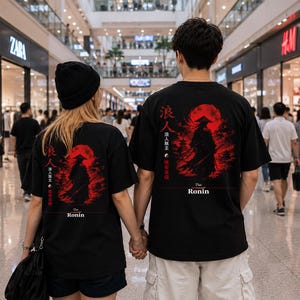 May include: Two black t-shirts featuring a graphic print on the back. The design displays a silhouette of a ronin warrior standing before a large red sun, accompanied by Japanese kanji characters and the text "The Ronin" in white lettering.