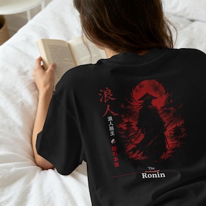 May include: A black t-shirt featuring a red graphic print on the back. The design depicts a silhouetted figure in a conical hat against a large red moon, accompanied by Japanese kanji characters, a yin-yang symbol, and the text The Ronin.