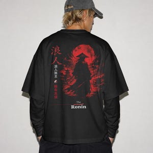 May include: A black long-sleeve t-shirt featuring a graphic print on the back. The design displays a silhouette of a samurai in a conical hat against a large red moon, with Japanese kanji characters, a yin-yang symbol, and the text The Ronin.