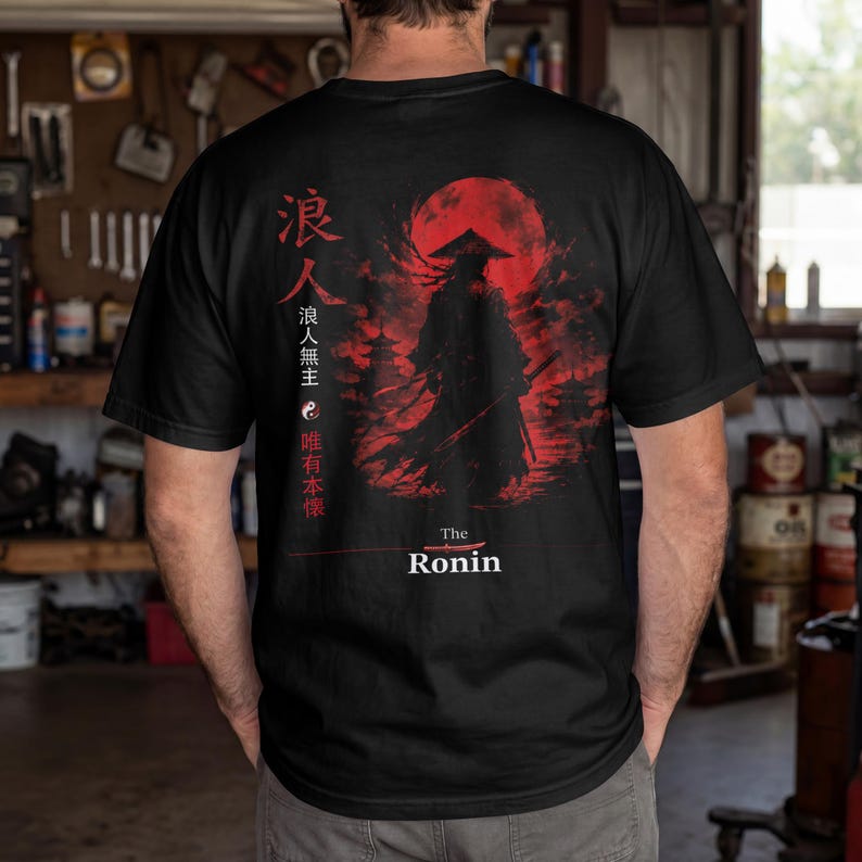 May include: A black t-shirt featuring a graphic print of a silhouetted samurai figure standing before a large red sun. The design includes Japanese kanji characters, a yin-yang symbol, and the text "The Ronin" printed in white at the bottom.