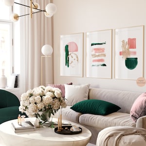 Könnte beinhalten: Drei abstrakte Kunstdrucke in goldenen Rahmen mit rosa, grünen und goldenen geometrischen Formen. Die Drucke hängen an einer weißen Wand über einem beigen Sofa mit grünen und rosa Kissen.