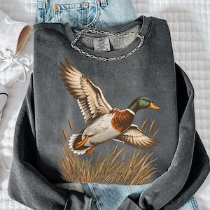 May include: A charcoal gray crewneck sweatshirt featuring a graphic of a mallard duck in flight over tall golden reeds. The sweatshirt is paired with light blue denim jeans and white high-top sneakers with white laces and rubber soles.
