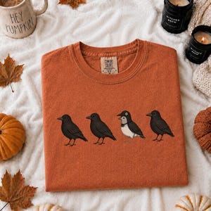 May include: A rust-colored crew neck t-shirt featuring a graphic of four birds standing in a row. Three of the birds are solid black, while the third bird from the left is black and white with a distinct white chest and a small white eye patch.
