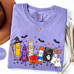May include: A violet short-sleeve t-shirt featuring a graphic print of anthropomorphic school supplies dressed in Halloween costumes, including a pencil witch, a crayon vampire, a mummy ruler, a pumpkin apple, a glue stick, and ghosts.