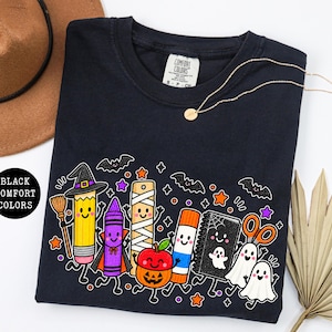 May include: A black t-shirt featuring a graphic print of cartoon school supplies dressed for Halloween, including a pencil with a witch hat, a crayon in a cape, a mummy ruler, a pumpkin apple, a glue stick, a notebook, and two ghosts.