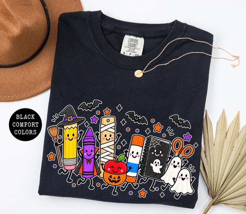 May include: A black t-shirt featuring a graphic print of cartoon school supplies dressed for Halloween, including a pencil with a witch hat, a crayon in a cape, a mummy ruler, a pumpkin apple, a glue stick, a notebook, and two ghosts.