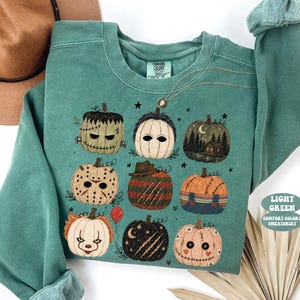 May include: A light green crewneck sweatshirt featuring a graphic print of nine stylized pumpkins decorated to resemble various horror movie characters. The design includes pumpkins with stitches, masks, and patterns, along with small stars and a red balloon.