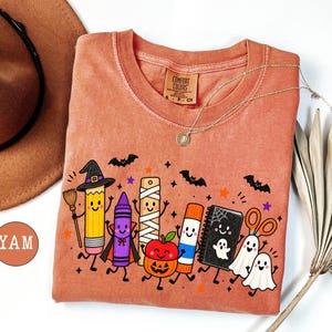 May include: A terracotta-colored t-shirt featuring a graphic print of anthropomorphic school supplies dressed for Halloween, including a pencil with a witch hat, a crayon, a mummy ruler, a pumpkin apple, a glue stick, a notebook, and ghosts.