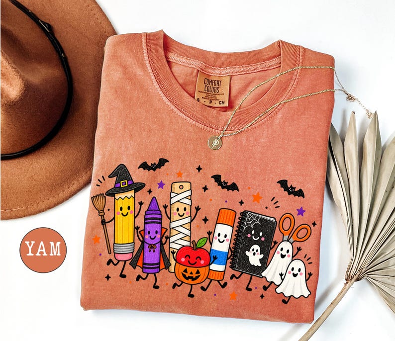 May include: A terracotta-colored t-shirt featuring a graphic print of anthropomorphic school supplies dressed for Halloween, including a pencil with a witch hat, a crayon, a mummy ruler, a pumpkin apple, a glue stick, a notebook, and ghosts.