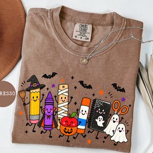 May include: An espresso-colored t-shirt featuring a graphic print of anthropomorphic school supplies dressed in Halloween costumes, including a pencil witch, a vampire crayon, a mummy ruler, a pumpkin apple, a glue stick, a ghost notebook, and scissors.