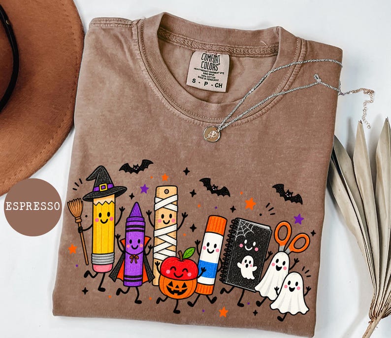 May include: An espresso-colored t-shirt featuring a graphic print of anthropomorphic school supplies dressed in Halloween costumes, including a pencil witch, a vampire crayon, a mummy ruler, a pumpkin apple, a glue stick, a ghost notebook, and scissors.