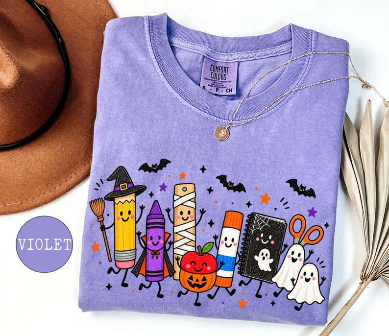 May include: A violet short-sleeve t-shirt featuring a graphic print of anthropomorphic school supplies dressed in Halloween costumes, including a pencil witch, a crayon vampire, a mummy ruler, a pumpkin apple, a glue stick, and ghosts.