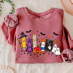 May include: A dusty rose long-sleeve sweatshirt featuring a graphic print of cartoon school supplies dressed for Halloween, including a pencil witch, a crayon vampire, a mummy ruler, a pumpkin, a glue stick, a notebook, and two ghosts.