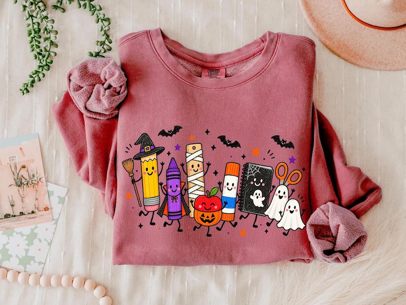 May include: A dusty rose long-sleeve sweatshirt featuring a graphic print of cartoon school supplies dressed for Halloween, including a pencil witch, a crayon vampire, a mummy ruler, a pumpkin, a glue stick, a notebook, and two ghosts.