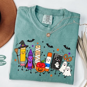 May include: A light green short-sleeve t-shirt featuring a graphic of cartoon school supplies dressed in Halloween costumes, including a pencil witch, a crayon vampire, a mummy ruler, a pumpkin apple, a glue stick, a ghost notebook, and ghosts.