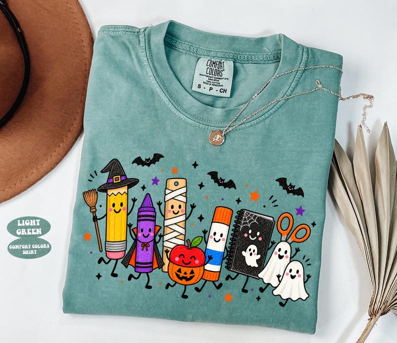 May include: A light green short-sleeve t-shirt featuring a graphic of cartoon school supplies dressed in Halloween costumes, including a pencil witch, a crayon vampire, a mummy ruler, a pumpkin apple, a glue stick, a ghost notebook, and ghosts.