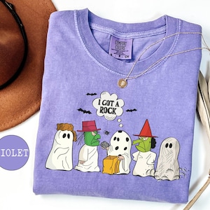 May include: A violet t-shirt featuring a graphic of five characters in ghost costumes, including two green-faced witches and one holding a rock with a speech bubble that says I GOT A ROCK. The shirt has a crew neck and a Comfort Colors brand label.