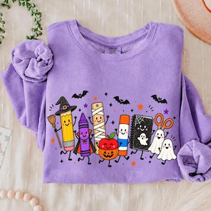 May include: A lavender crewneck sweatshirt featuring a graphic print of anthropomorphic school supplies dressed for Halloween, including a pencil with a witch hat, a crayon vampire, a mummy ruler, a jack-o'-lantern apple, a glue stick, and ghosts.