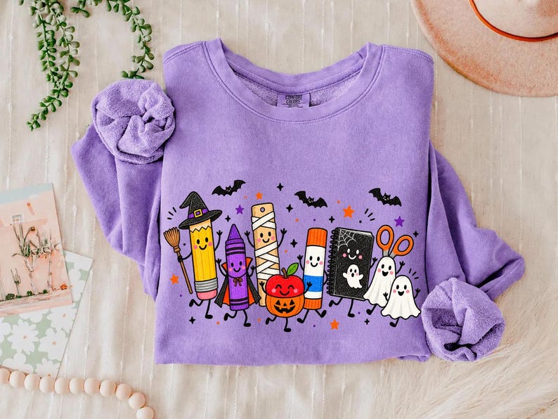 May include: A lavender crewneck sweatshirt featuring a graphic print of anthropomorphic school supplies dressed for Halloween, including a pencil with a witch hat, a crayon vampire, a mummy ruler, a jack-o'-lantern apple, a glue stick, and ghosts.