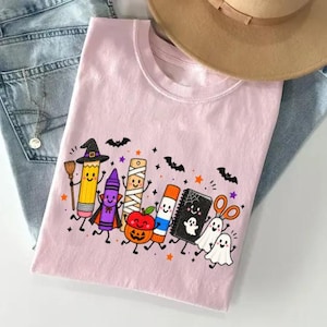 May include: A light pink t-shirt featuring a graphic print of anthropomorphic school supplies dressed for Halloween, including a pencil with a witch hat, a crayon vampire, a mummy ruler, a jack-o'-lantern apple, a glue stick, and two ghosts.