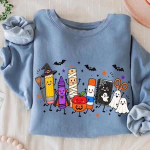 May include: A dusty blue crewneck sweatshirt featuring a colorful graphic of anthropomorphic school supplies dressed in Halloween costumes, including a pencil witch, a crayon vampire, a mummy ruler, a pumpkin, a glue stick, a notebook, and ghosts.