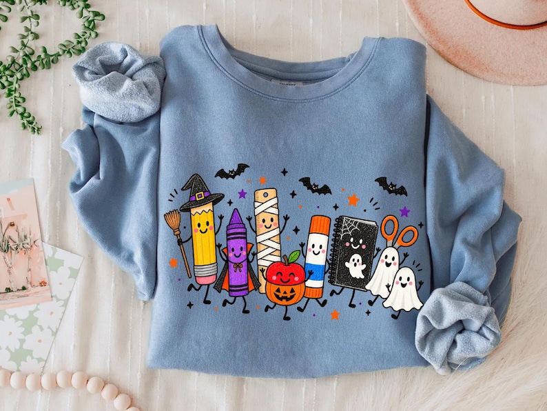May include: A dusty blue crewneck sweatshirt featuring a colorful graphic of anthropomorphic school supplies dressed in Halloween costumes, including a pencil witch, a crayon vampire, a mummy ruler, a pumpkin, a glue stick, a notebook, and ghosts.