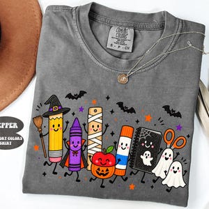 May include: A pepper-colored Comfort Colors t-shirt featuring a graphic of cartoon school supplies dressed in Halloween costumes, including a pencil witch, a crayon vampire, a mummy ruler, a pumpkin apple, a glue stick, a ghost notebook, and ghosts.