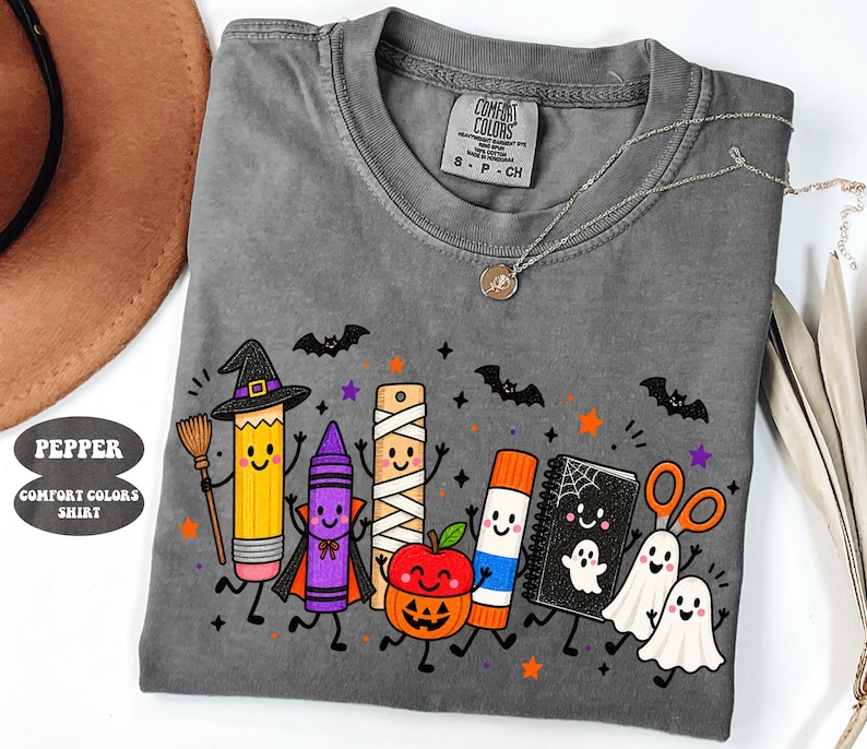 May include: A pepper-colored Comfort Colors t-shirt featuring a graphic of cartoon school supplies dressed in Halloween costumes, including a pencil witch, a crayon vampire, a mummy ruler, a pumpkin apple, a glue stick, a ghost notebook, and ghosts.