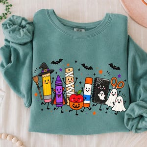 May include: A teal crewneck sweatshirt featuring a graphic print of anthropomorphic school supplies dressed for Halloween, including a pencil witch, a crayon vampire, a mummy ruler, a pumpkin, a glue stick, a notebook, and ghost scissors.