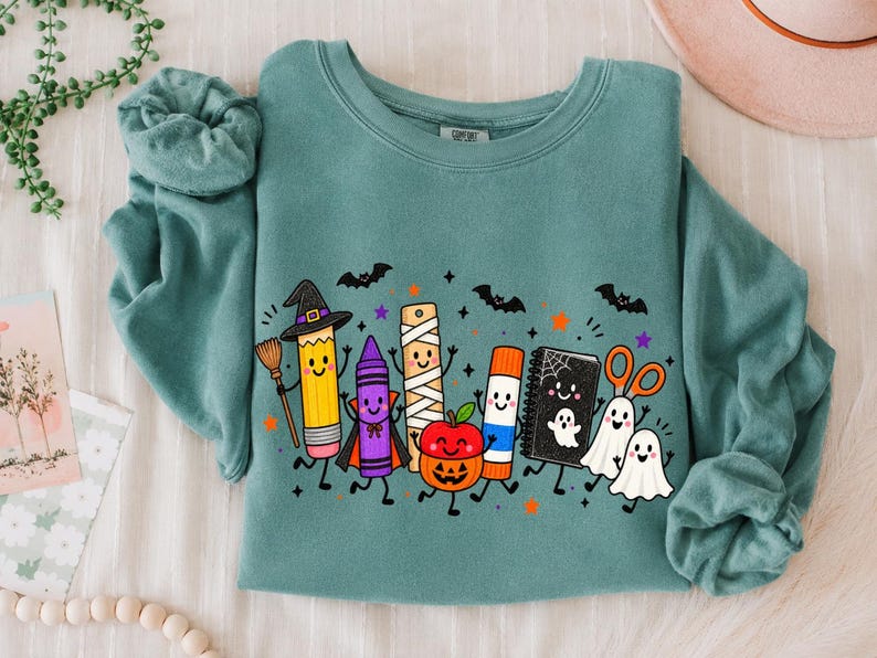 May include: A teal crewneck sweatshirt featuring a graphic print of anthropomorphic school supplies dressed for Halloween, including a pencil witch, a crayon vampire, a mummy ruler, a pumpkin, a glue stick, a notebook, and ghost scissors.