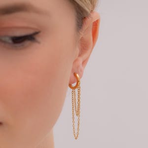 May include: Gold-colored hoop earring with dangling chain accents. The earring is a half-circle design with multiple delicate chains hanging from the bottom. The jewelry is shown against a neutral background.