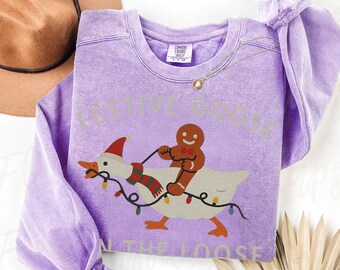 May include: A light purple sweatshirt with the text "FESTIVE GOOSE ON THE LOOSE". The graphic features a goose wearing a Santa hat and scarf, ridden by a gingerbread man, with Christmas lights.