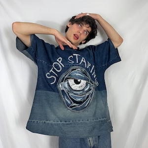 Op de afbeelding: Een blauw denim shirt met een grote oogprint en de tekst "STOP STARING" in wit. Het shirt is gemaakt van distressed denim stof en heeft een relaxed fit.