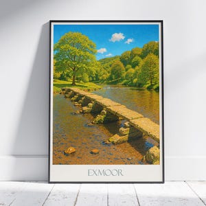Puede incluir: Una impresión enmarcada de un paisaje pintoresco con un puente de piedra que cruza un río. La escena presenta exuberantes árboles verdes y un cielo azul claro con nubes. La palabra "EXMOOR" está impresa en la parte inferior.