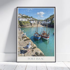 May include: A framed print of Port Isaac, featuring two sailboats in a harbor with turquoise water. White buildings line the hillside under a blue sky. Seagulls perch on a stone wall in the foreground. The text "PORT ISAAC" is at the bottom.