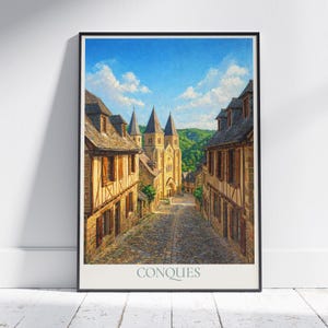 May include: Framed print of a European village street scene. The artwork depicts a cobblestone street leading to a church with multiple spires. Buildings with brown roofs and wooden shutters line the street. The sky is blue with fluffy white clouds. The word "CONQUES" is printed at the bottom.