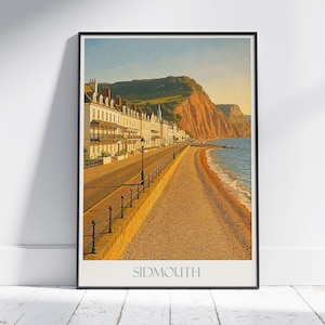 May include: Framed Sidmouth print featuring a coastal scene with a pebble beach, road, and buildings. The artwork showcases a vibrant landscape with a mountain in the background and the text "SIDMOUTH" at the bottom.