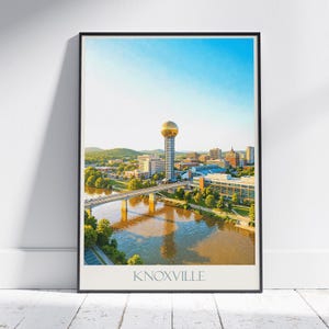 Puede incluir: Una impresión enmarcada que muestra una vista aérea de Knoxville, Tennessee, con la Sunsphere. La imagen incluye un río, un puente, edificios y un cielo azul claro. La palabra "KNOXVILLE" está impresa en la parte inferior.