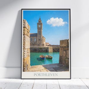 May include: Framed print of Porthleven, featuring a stone clock tower and harbor with two small boats. The image is framed by stone walls, with the town's name printed at the bottom. The sky is blue with a single cloud.