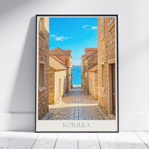 May include: A framed print of a narrow cobblestone street leading to the sea. The buildings are made of light brown stone with red tile roofs. The sky is bright blue, and the sea is visible at the end of the street. The word "KORČULA" is printed at the bottom.