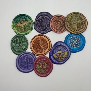 May include: A collection of colorful wax seals with various designs, including a dragon, a compass rose, a ship, and a die. The seals are in shades of green, blue, purple, gold, and red, and are arranged on a white surface.
