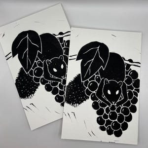 May include: Two black and white linocut prints. Each print features a squirrel holding grapes, with large leaves and a vine. The squirrel and grapes are black, and the background is white. The prints are on white paper.