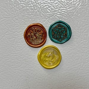 May include: Three wax seals in copper, teal, and yellow. The copper seal depicts a ship, the teal seal features a twenty-sided die, and the yellow seal shows a dragon. These seals are likely used for decorative purposes.