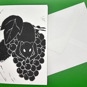 May include: Black and white art print of a mouse holding grapes. The illustration style is simple, with bold lines and a minimalist aesthetic. A white envelope is next to the card. The background is green.