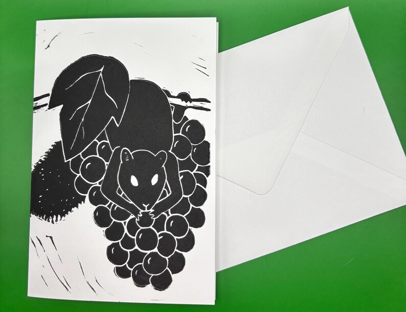 May include: Black and white art print of a mouse holding grapes. The illustration style is simple, with bold lines and a minimalist aesthetic. A white envelope is next to the card. The background is green.