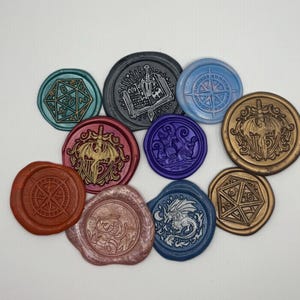 May include: A collection of colorful wax seals with various designs, including a compass, a dragon, a d20 die, and a sword and shield. The seals are in shades of blue, red, gold, and purple, and are arranged on a white surface.