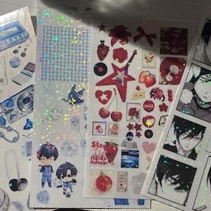 Può includere: Una collezione di adesivi decorativi con vari disegni. Gli adesivi includono immagini di personaggi anime, strumenti musicali, cibo e dispositivi elettronici. Gli adesivi sono in tonalità blu, rosse e bianche, con una finitura glitterata.