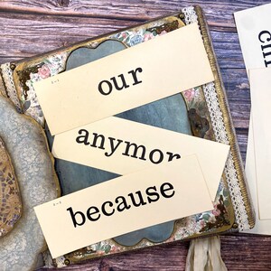 May include: Three beige paper cards with black text. The top card says "our", the middle card says "anymore", and the bottom card says "because". The cards are arranged on top of a vintage-style scrapbook with floral and lace embellishments.