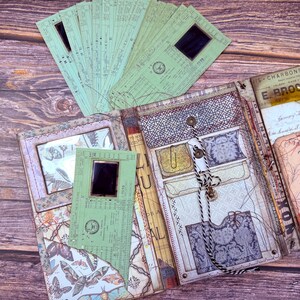 May include: A handcrafted journal with a vintage design. The journal includes patterned papers, pockets, and a button closure. Several green tickets are arranged around the journal, enhancing the antique theme. The journal is made with a variety of materials.