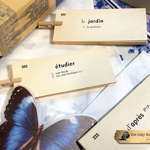 May include: A set of flashcards with French vocabulary words and definitions. The cards are beige with black text and are held together with wooden clothespins. The cards are on a white surface with a blue and white patterned background.